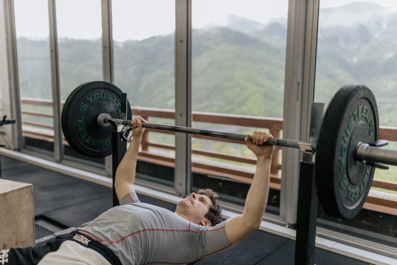 A man performs a bench press in a gym with mountain views, emphasizing strength and fitness.